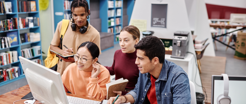A group of four young adults collaborating around a computer in a library, with bookshelves and a coffee machine in the background symbolizing 'Zamit student skill zone'.